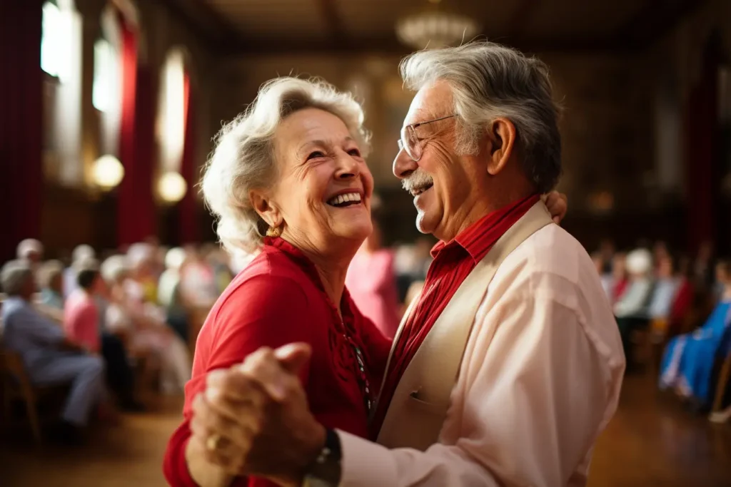 A joyful elderly couple dancing together in a ballroom, smiling warmly at each other. The woman wears a red dress and has short white hair, while the man wears glasses, a red shirt, and a white jacket. Other guests are seated in the blurred background of the elegant hall with tall windows and red curtains.