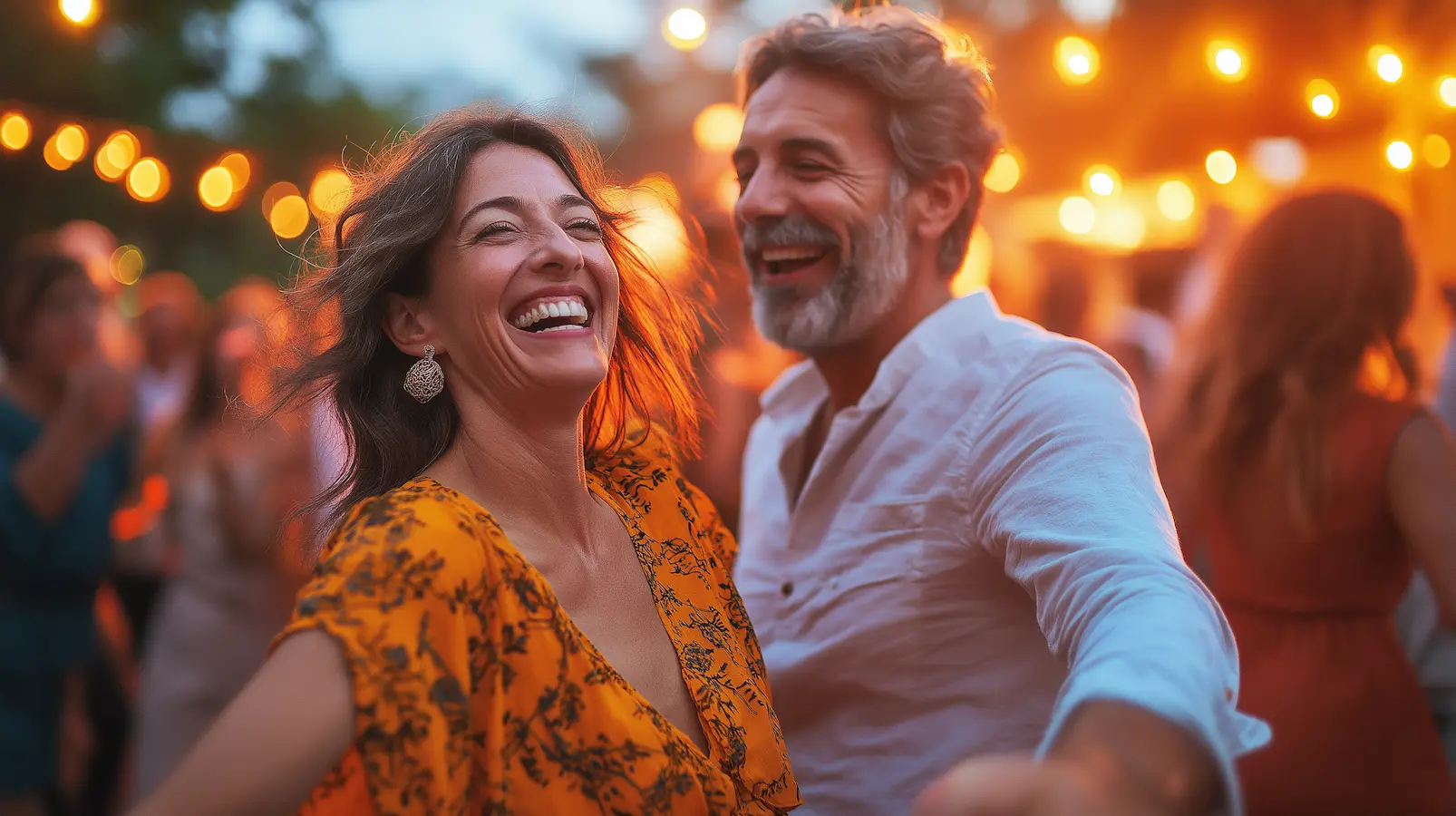 A joyful middle-aged couple laughing and dancing together at an outdoor evening event, with warm string lights glowing in the background. The woman wears a yellow floral dress and dangling earrings, while the man has a gray beard and white button-up shirt. Other guests are visible in the soft-focus background.