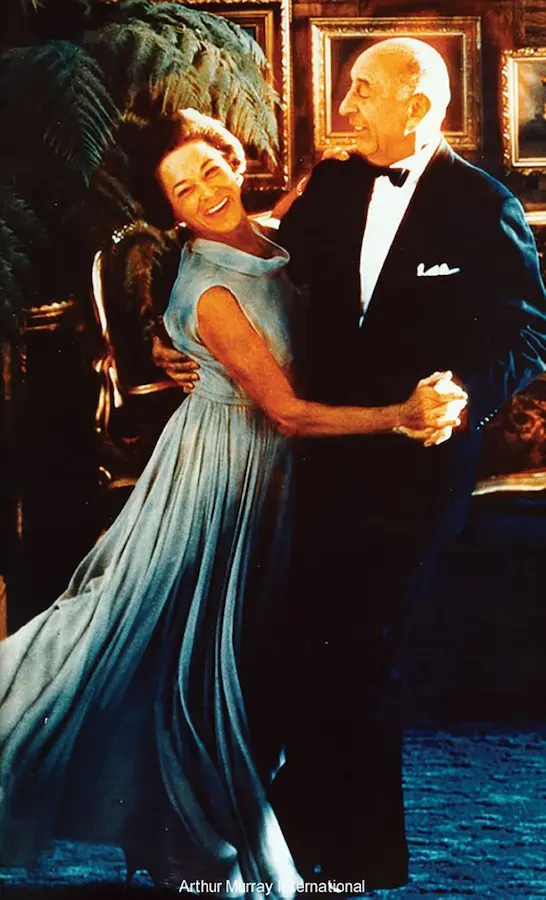 Arthur and Kathryn Murray dancing together in an elegant setting, with Kathryn wearing a flowing light blue gown and Arthur in a dark tuxedo with bow tie and pocket square. Both are smiling joyfully as they dance, with framed paintings and tropical plants visible in the background. Photo credited to Arthur Murray International.