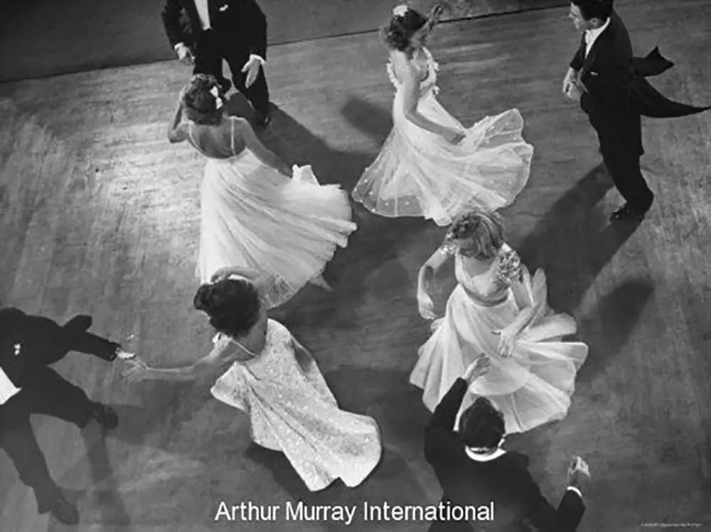 Vintage black and white overhead photograph of couples ballroom dancing on a wooden dance floor, with women wearing flowing white gowns and men in dark formal tuxedos, captured during an Arthur Murray International event.