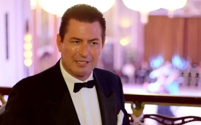 A middle-aged man wearing a formal black tuxedo with a white dress shirt and black bow tie, photographed at what appears to be an elegant event or banquet hall with soft, blurred lighting and chandeliers visible in the background. He has dark hair and is looking slightly to the side with a composed expression.