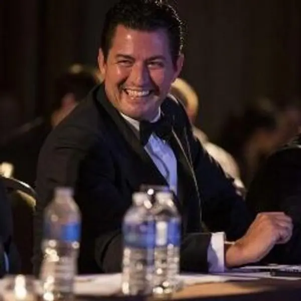 A smiling man in a dark suit and light blue dress shirt seated at a conference or event table with water bottles and papers in front of him, with a dark background suggesting a formal indoor setting.