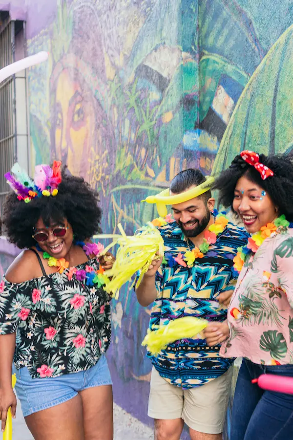 Three friends laughing and celebrating together in front of a colorful street mural. They are dressed in vibrant, festive outfits with tropical prints, flower leis, and colorful accessories. A woman on the left wears a floral top, denim shorts, sunglasses, and a multicolored feather headband. A bearded man in the center wears a blue patterned shirt, a yellow hat, and a flower lei while holding yellow feathers. A woman on the right wears a pink tropical blouse, a red polka-dot headband, and face glitter, all enjoying a lively, carnival-like atmosphere.