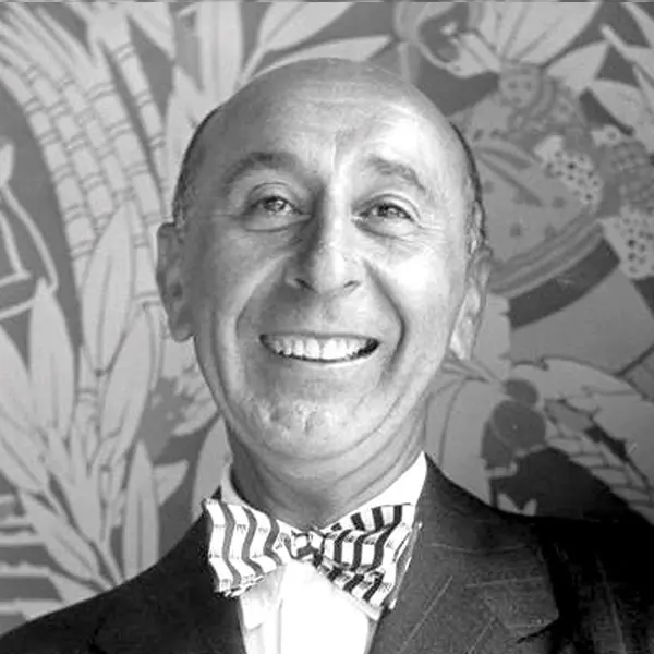Black and white portrait of Arthur, a bald middle-aged man smiling broadly, wearing a dark suit jacket, striped dress shirt, and a striped bow tie, posed in front of a colorful floral patterned wallpaper or mural background.