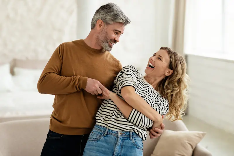 A happy middle-aged couple dancing and laughing together in their bright living room, with the man in a brown sweater holding the woman in a black and white striped top as she leans back with joy.