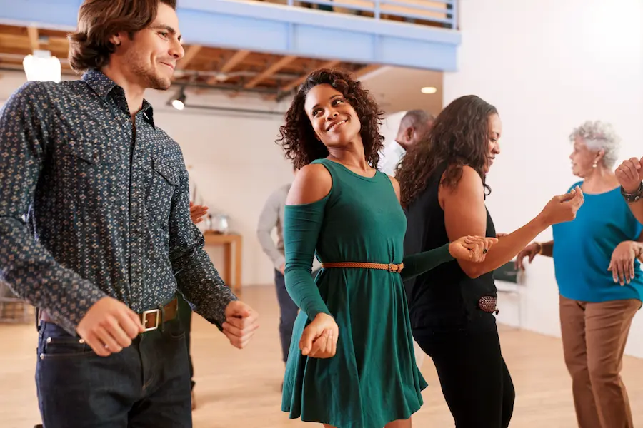 Couples of various ages smiling and dancing together in a bright dance studio during a group dance class, with a young woman in a teal dress and a man in a patterned shirt featured prominently in the foreground.
