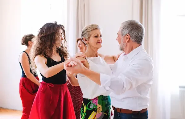 A group of adults participating in a dance lesson in a bright studio. In the foreground, a young woman with curly brown hair wearing a black top and red skirt dances with a middle-aged couple, while a blonde woman in a floral skirt partners with a gray-haired man in a white shirt. Other participants can be seen practicing in the background.